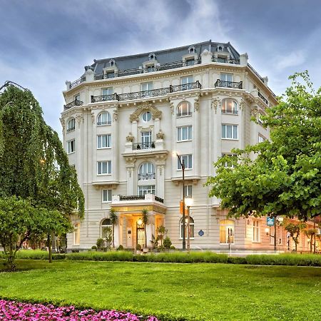 Hotel Carlton Bilbao Exterior photo The photo depicts a beautiful, historic building, likely a hotel or a residence, featuring an elegant architectural style with white facade and ornate details. It is surrounded by a lush green lawn, trees, and colorful flower beds that enhance the ae