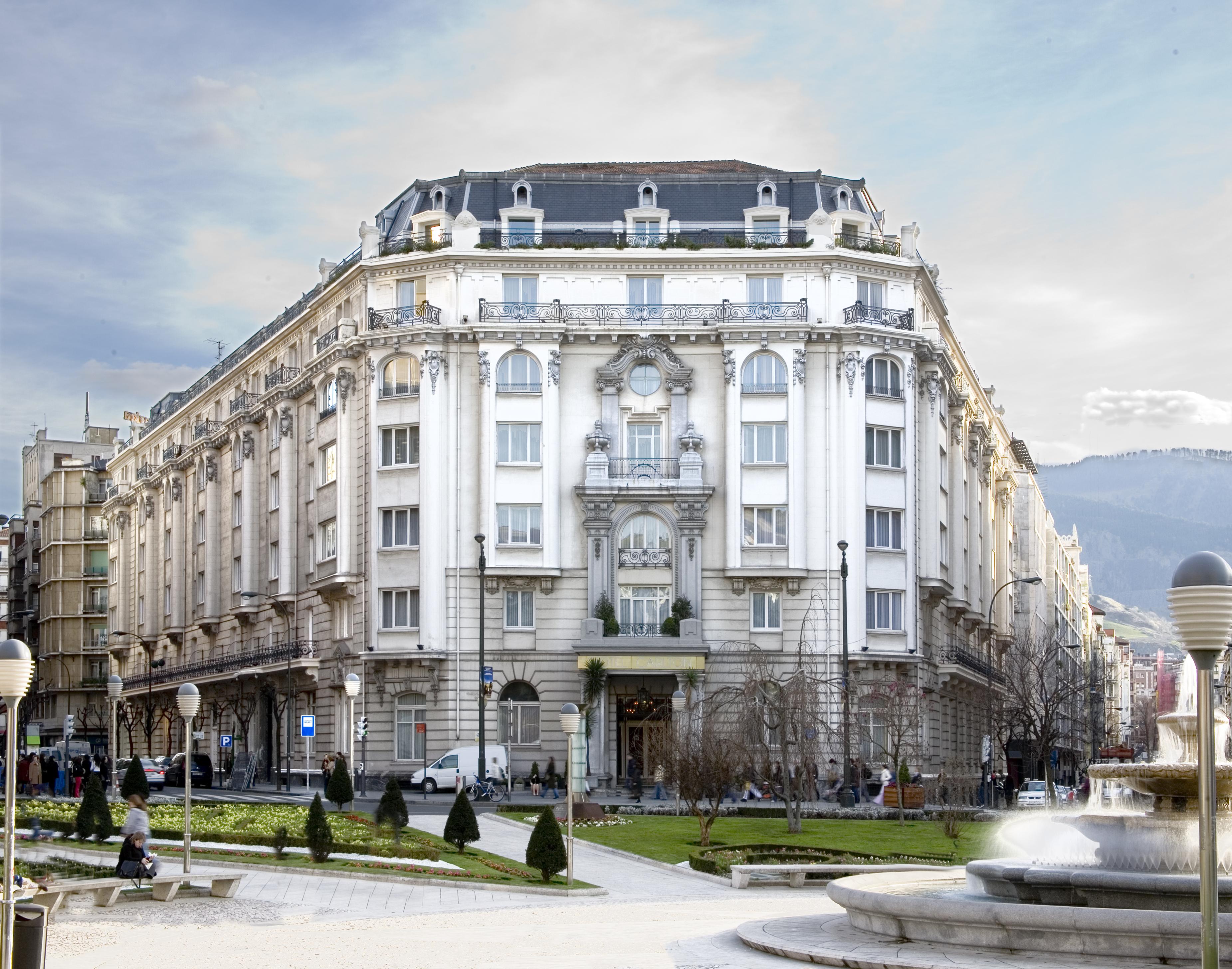 Hotel Carlton Bilbao Exterior photo The photo shows a grand, historical building with an ornate facade. The architecture features decorative elements, large windows, and a prominent entrance. Surrounding the building is a green area with well-maintained grass and small trees, creating 